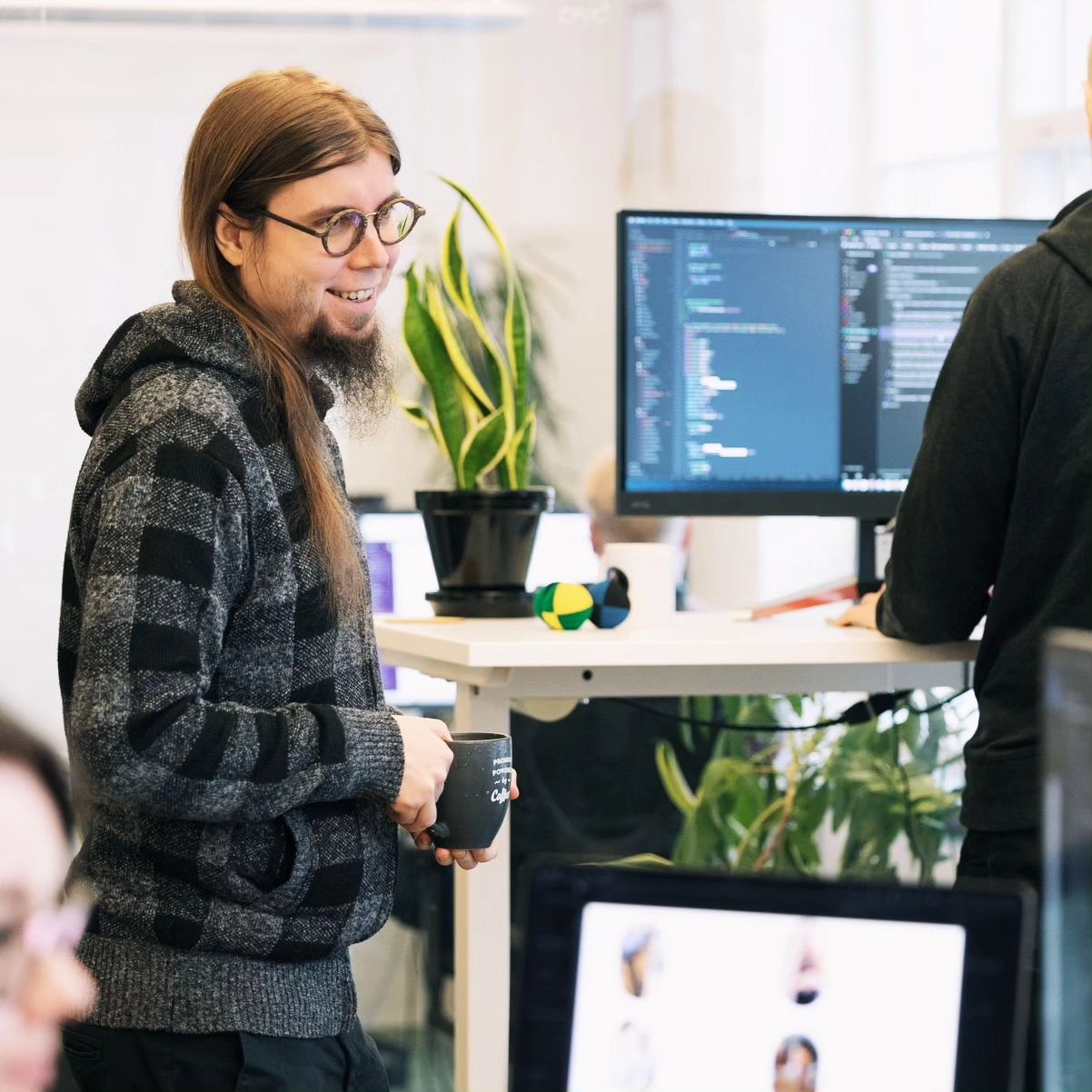 A person smiling with coffee cup in his hand, long haired, office full of plants, people working.